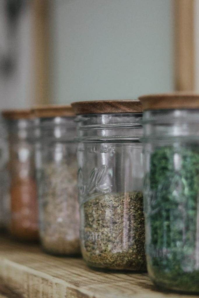 a row of pantry staples of herbs and spices in glass jars with wooden lids