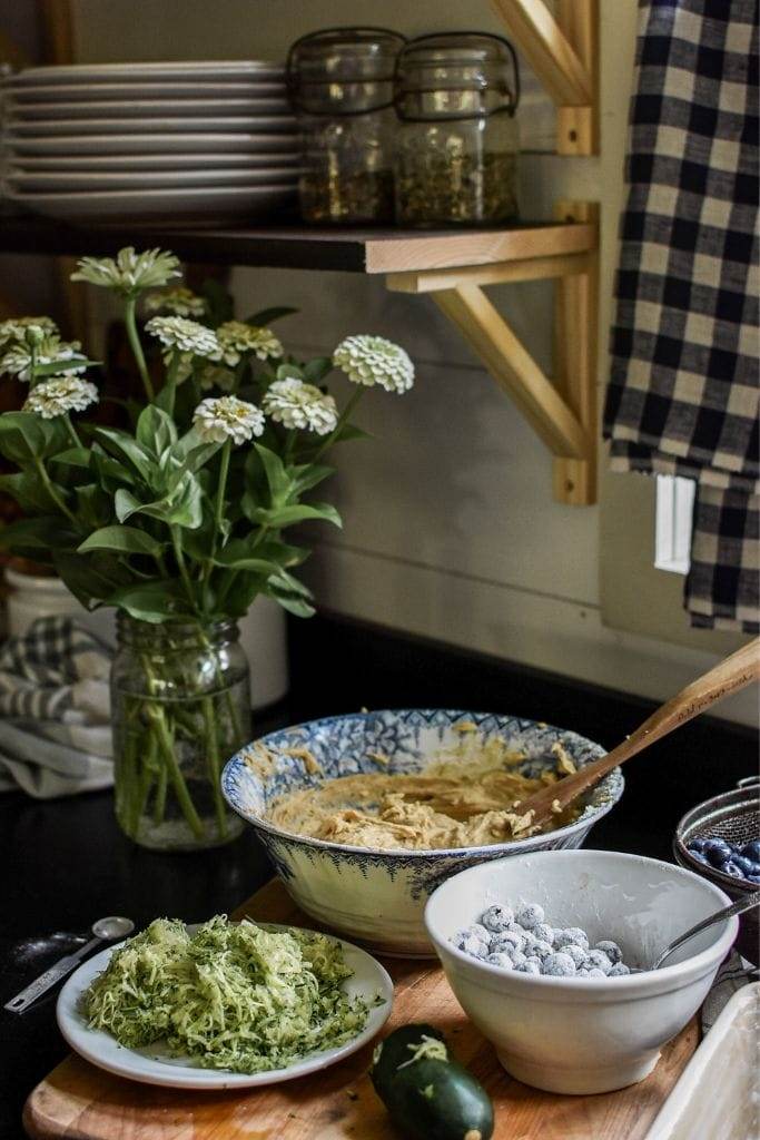 three bowls sitting on the counter. One has shredded zucchini, the other one has the batter for zucchini bread and one bowl has blueberries.