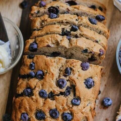 A loaf of zucchini blueberry bread partially sliced on a wooden cutting board with fresh blueberries nearby.