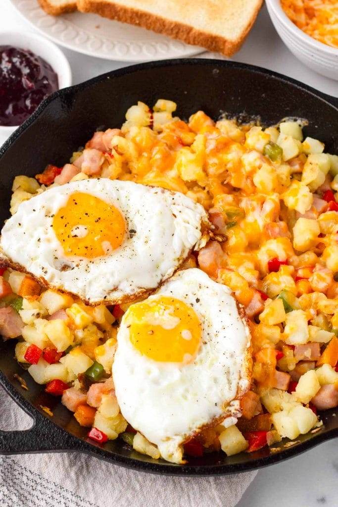 A skillet filled with a breakfast hash of scrambled eggs, diced ham, cubed hash brown potatoes, and sliced green peppers