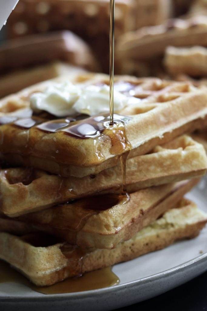 A close-up of a stack of three golden brown waffles, with a pat of melting butter on top and drizzled with syrup.