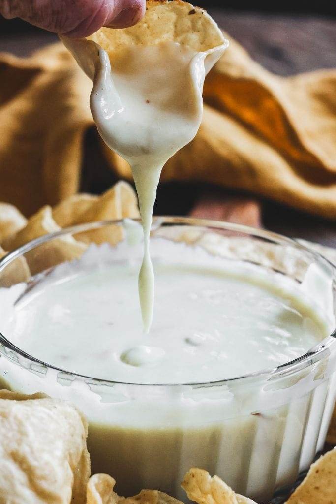 A bowl of white queso with a chip held up that has the queso dripping off of it.