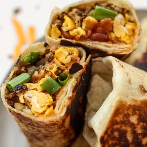 Close-up of a breakfast burrito cut in half, showcasing the crispy bacon, scrambled eggs, melted cheese, and fresh green onions inside.