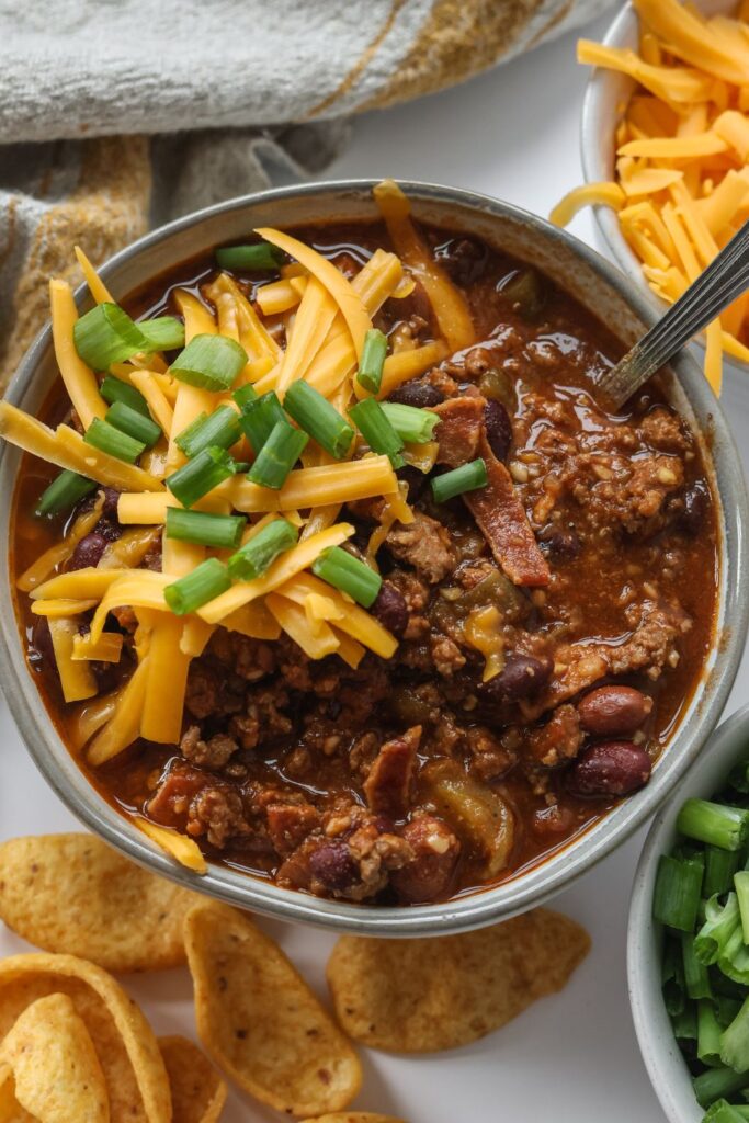 Close-up of a cozy bowl of bacon chili, topped with cheese, and chopped green onions.