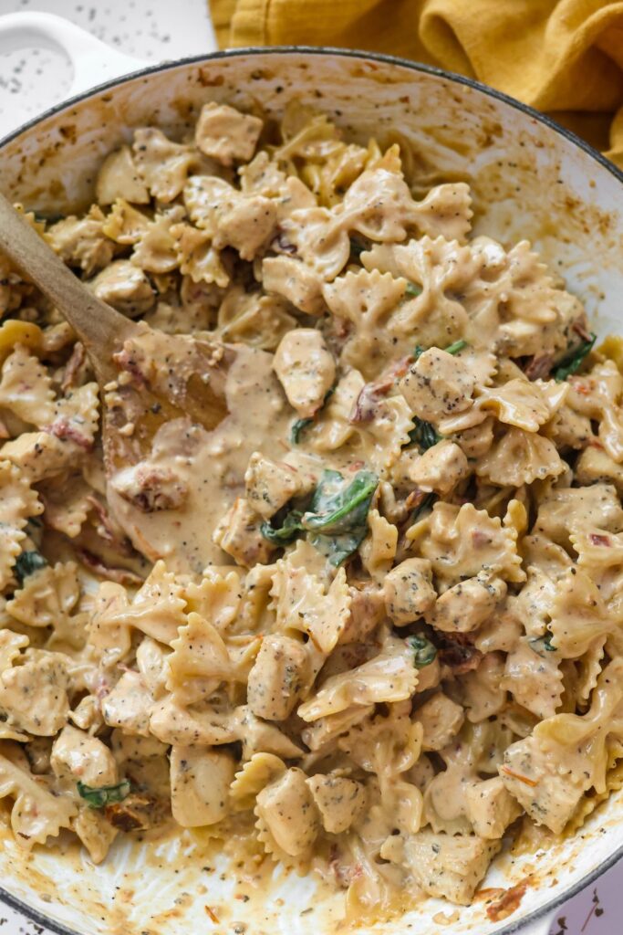 A serving spoon lifting a creamy, sun-dried tomato and spinach-filled Tuscan chicken pasta from a skillet