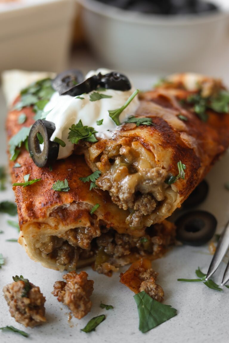 an upclose photo showing the creamy ground beef inside of an enchilada. it has sour cream and black olives on top of the enchilada