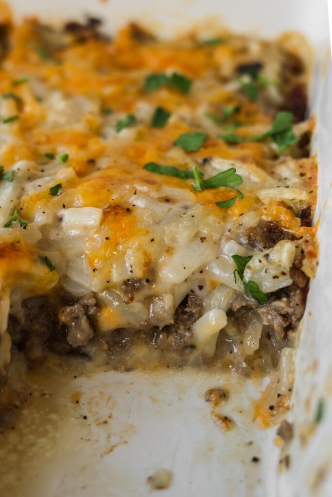 Baked hamburger hashbrown casserole in a dish, with a section cut out to reveal the creamy layers of ground beef, hashbrowns, and melted cheddar cheese.